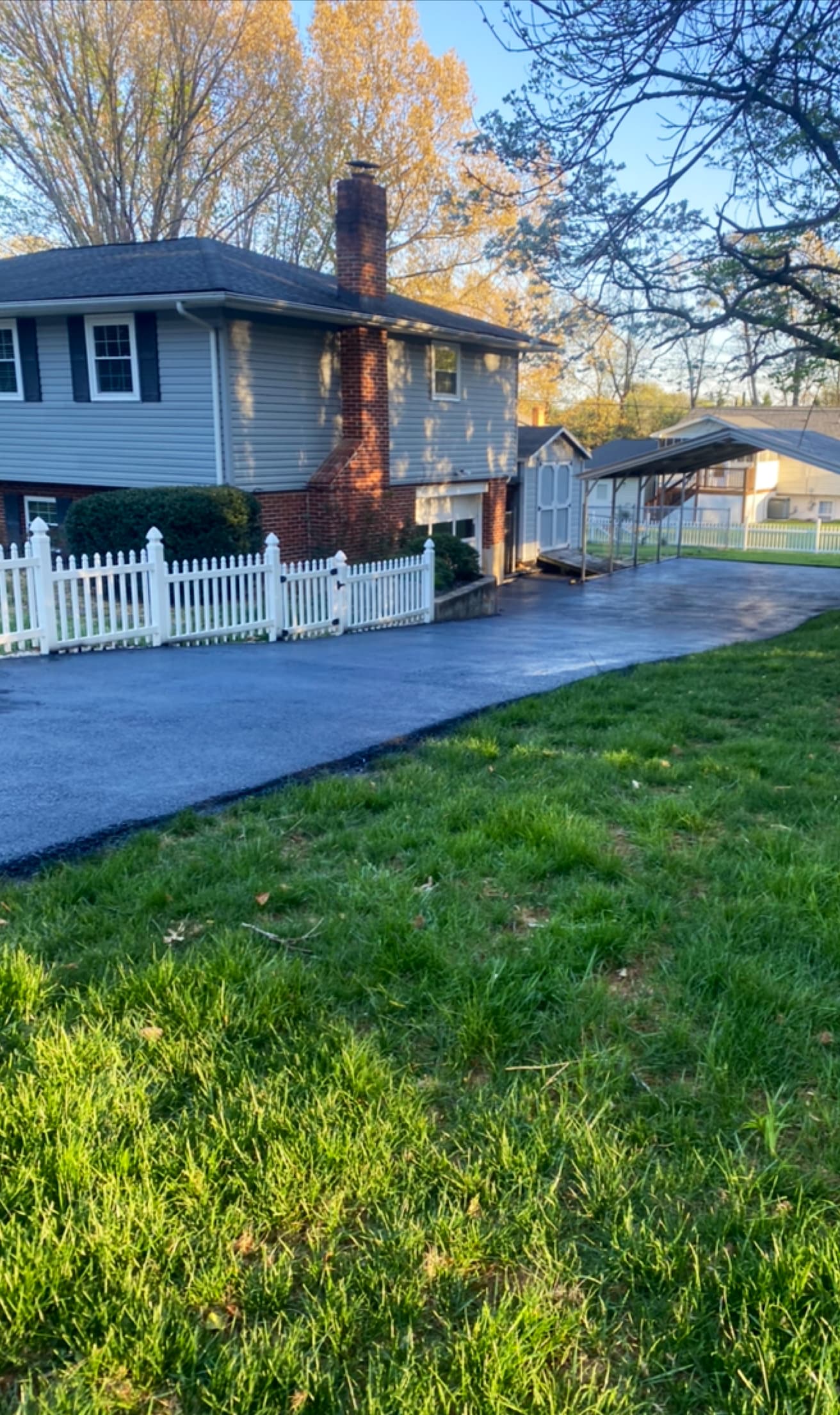 Freshly sealed black asphalt driveway beside a white picket fence and a gray house.