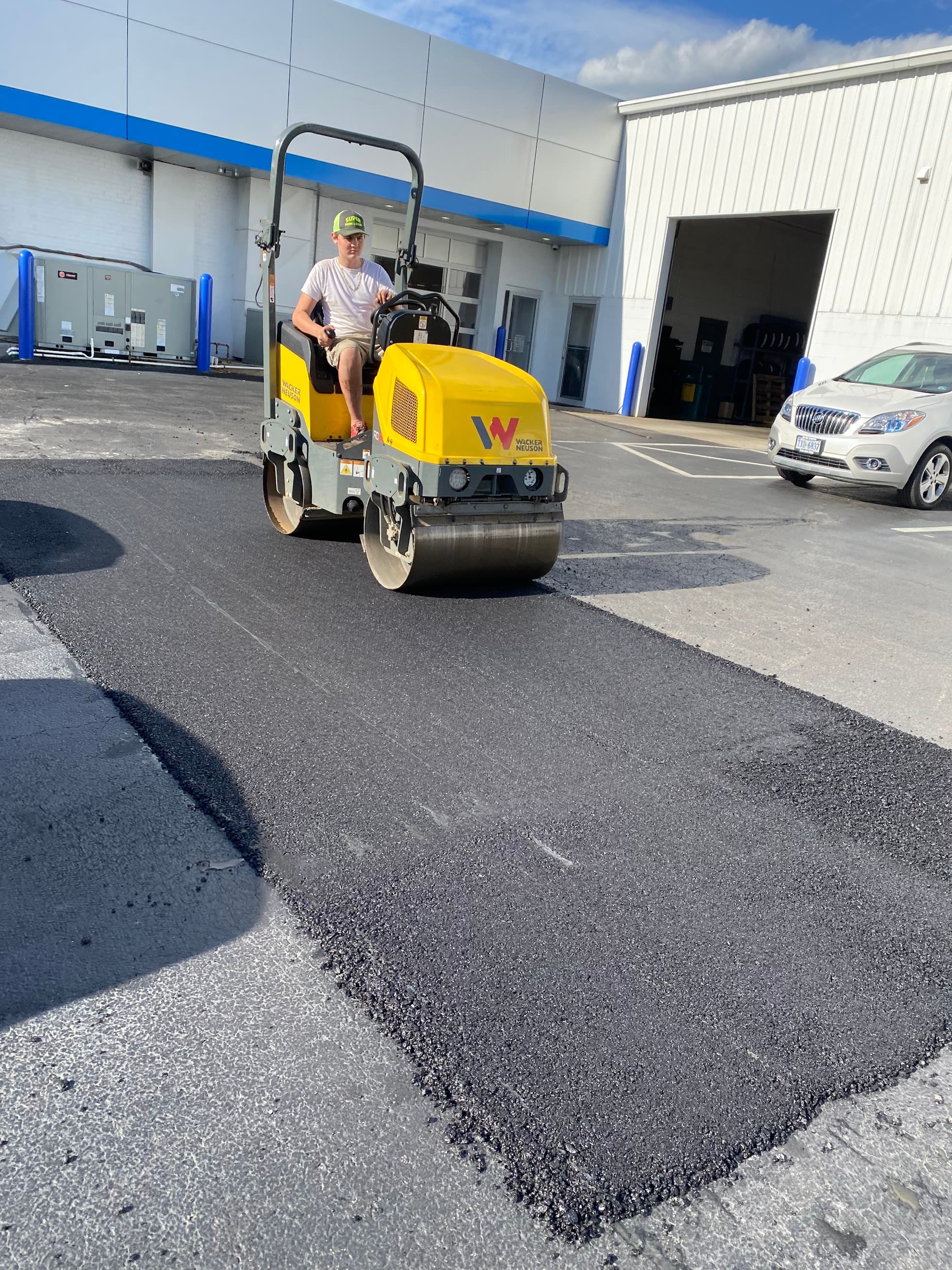 Worker operating a yellow asphalt roller to smooth a fresh patch of black pavement.