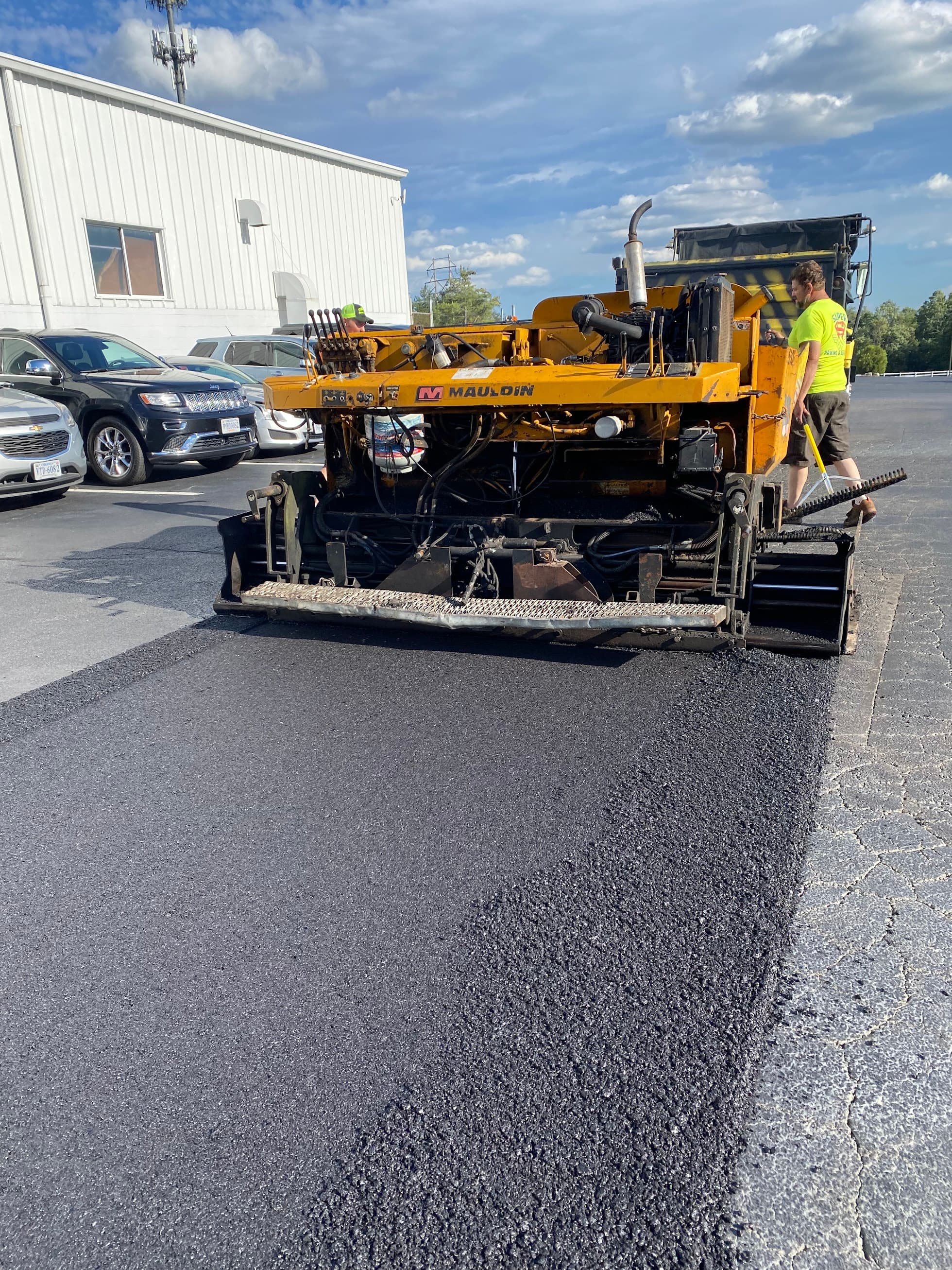 Yellow Mauldin paver laying fresh black asphalt in a parking lot next to parked cars.