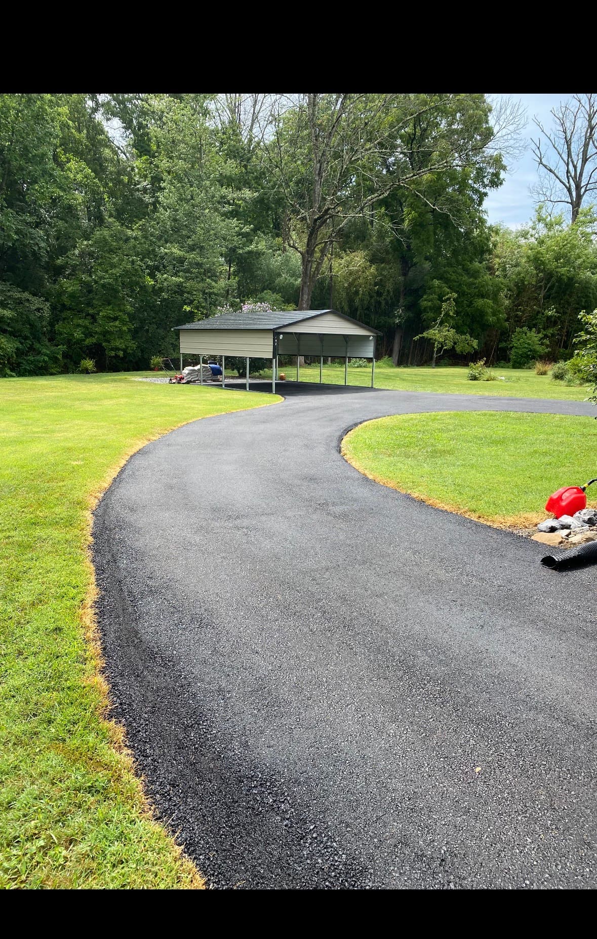 A newly paved asphalt driveway curves through a green lawn toward a metal carport.