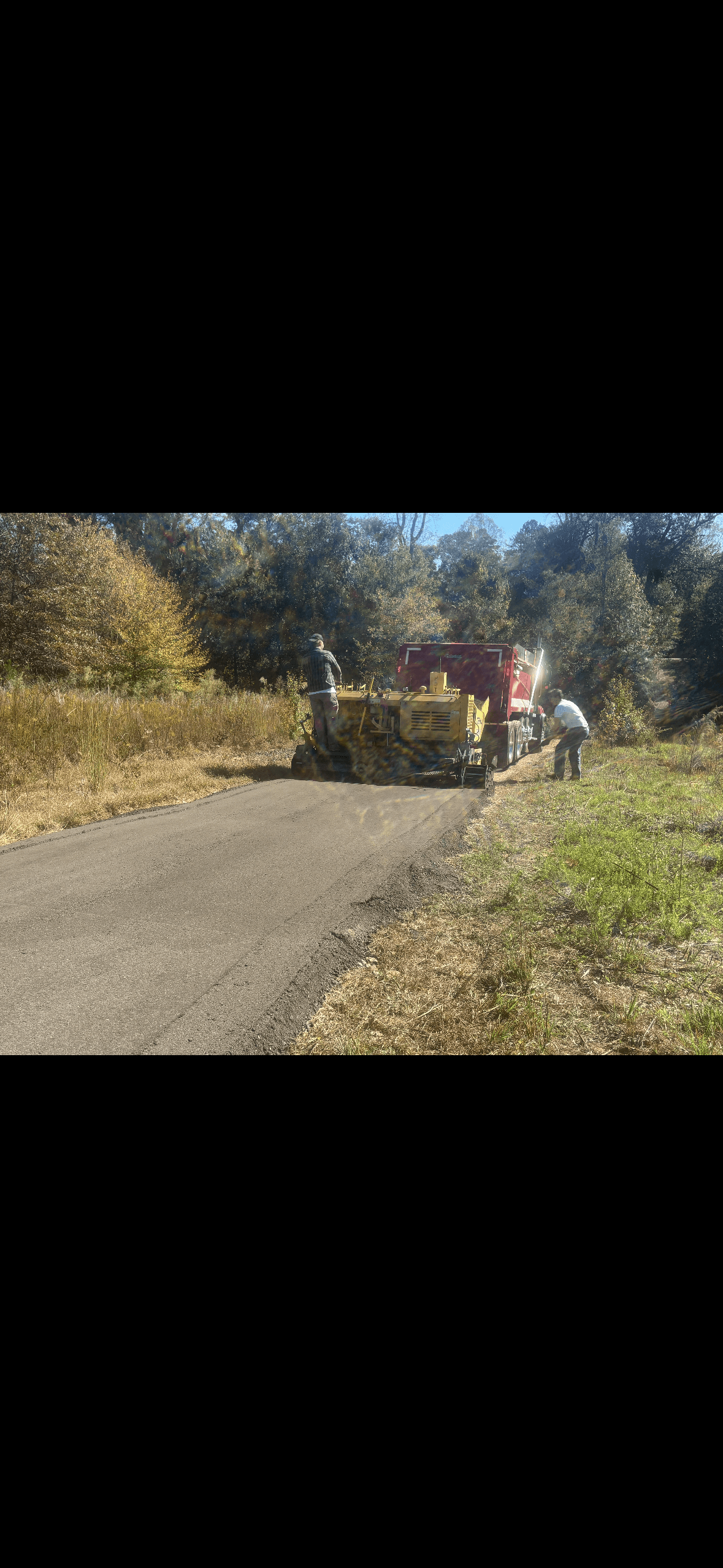 A yellow paver and red truck laying fresh asphalt on a narrow road through trees.