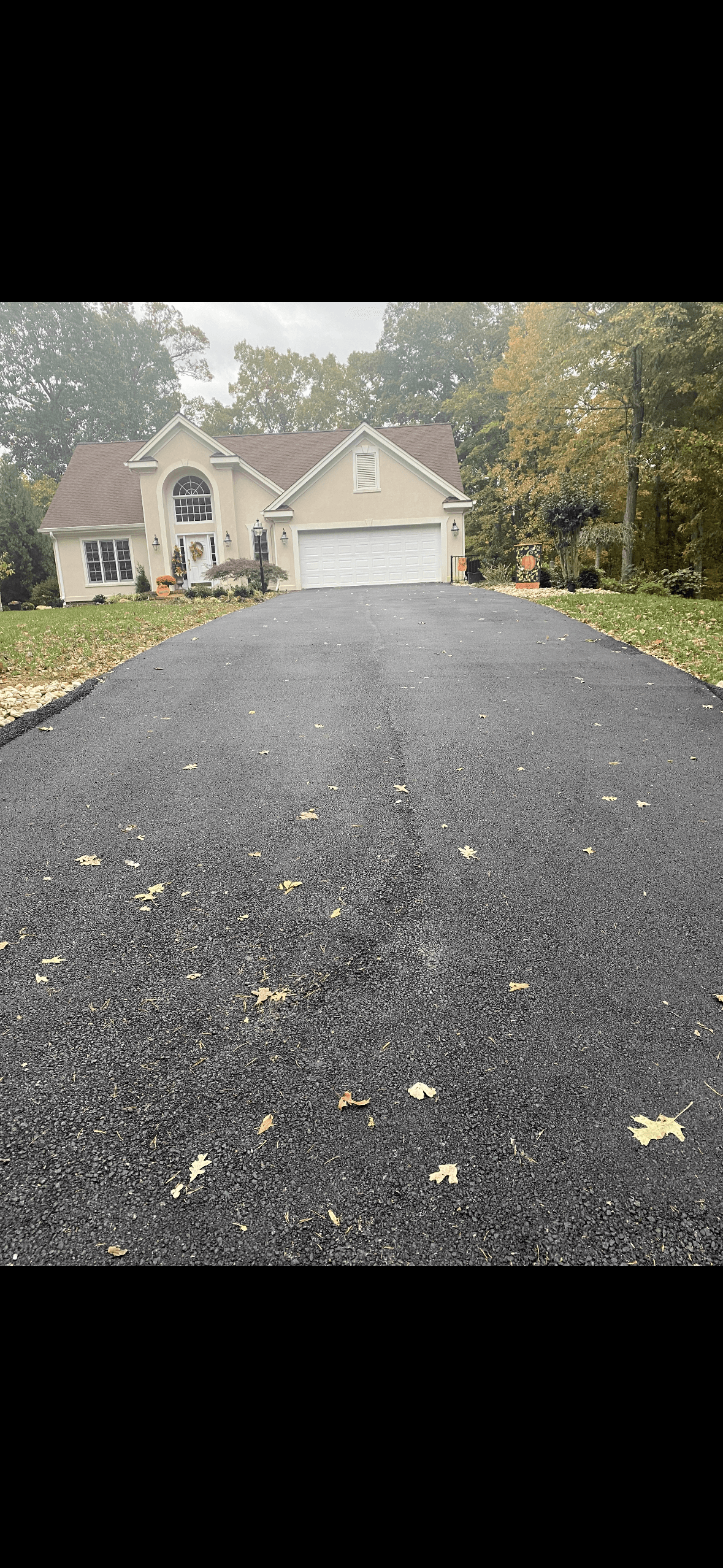 Long black asphalt driveway with scattered leaves leading to a beige house and white garage.